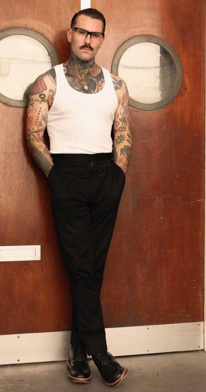 a man with tattoos standing in front of a door wearing black pants and a white tank top