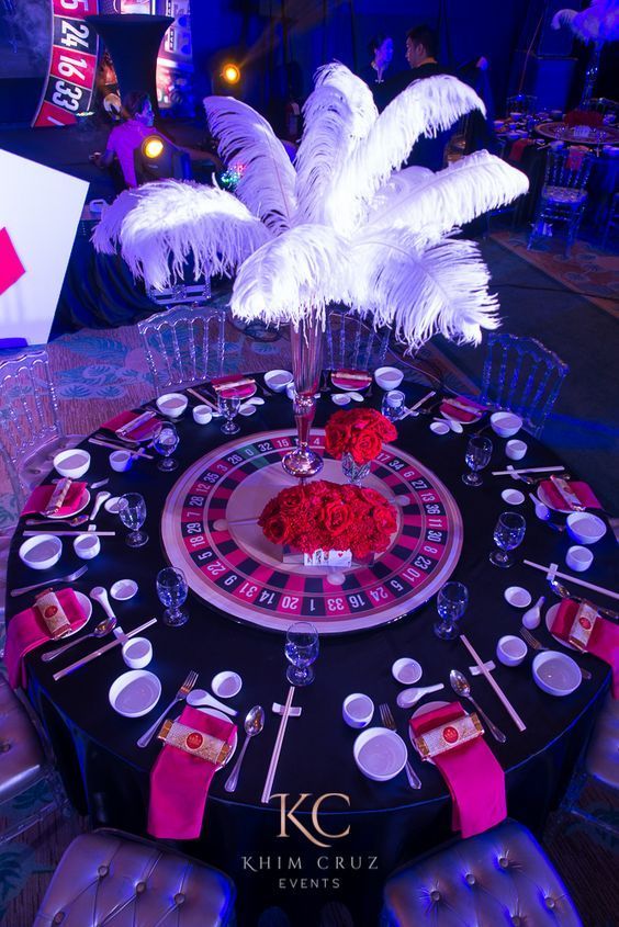 a casino roule with white feathers and red roses on the center table for an event