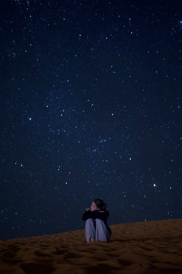 a man sitting on top of a sandy beach under a night sky filled with stars