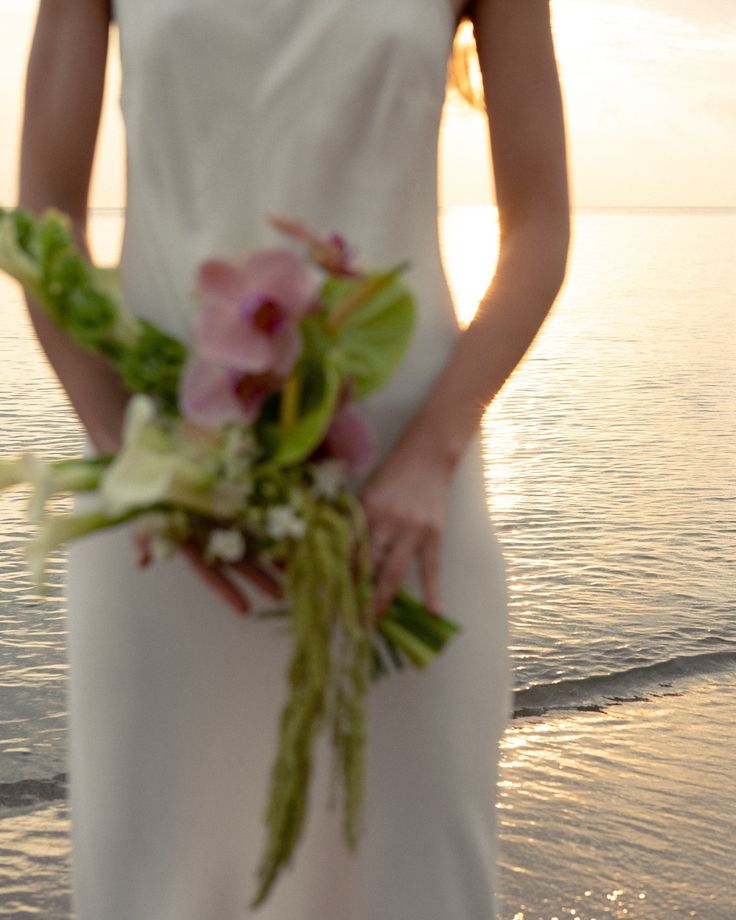 a woman in a white dress is holding a bouquet on the beach at sunset or sunrise