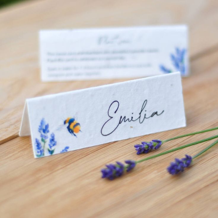 two place cards sitting on top of a wooden table next to lavender flowers and a bee