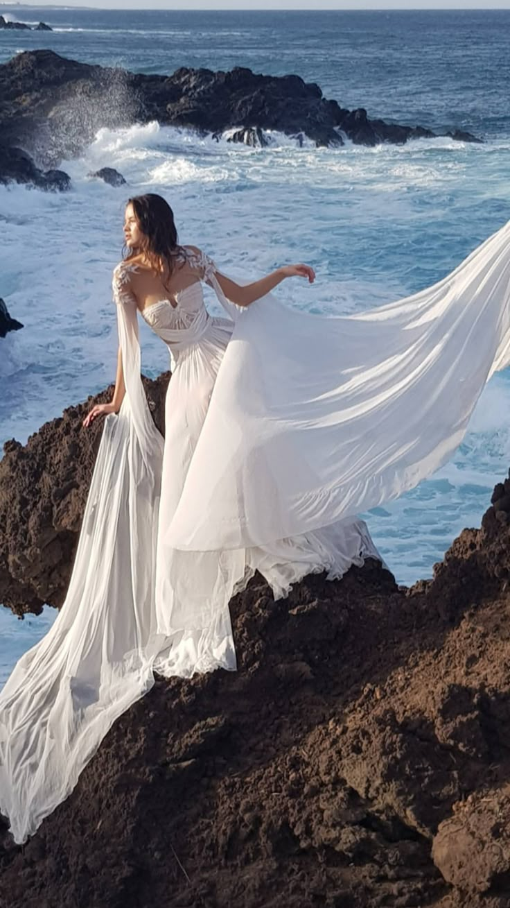 a woman standing on top of a rocky beach next to the ocean wearing a white dress