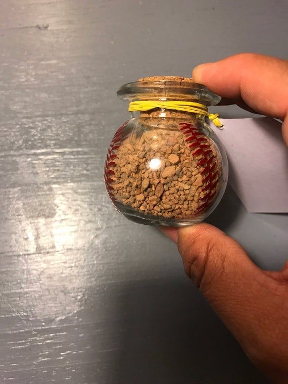 a person holding a baseball in a glass jar filled with dirt and yellow ribbon around it