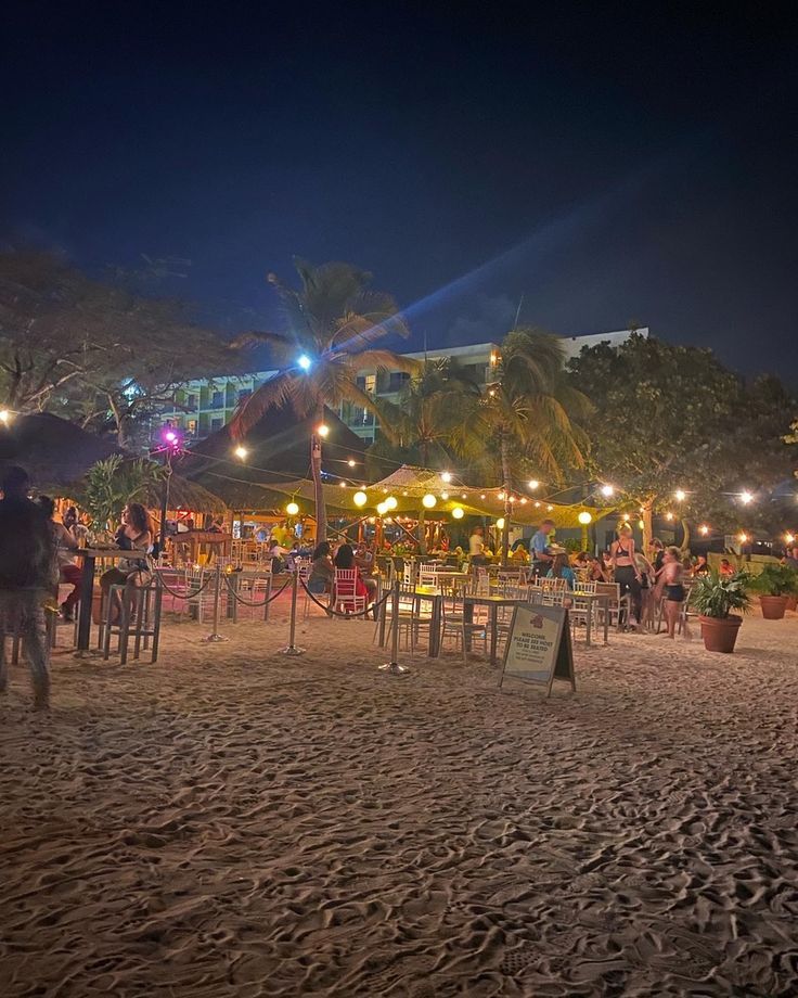 people are sitting at tables on the beach in front of an outdoor bar and restaurant