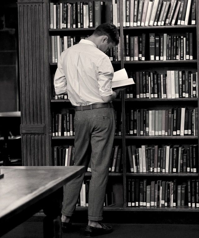 a man standing in front of a bookshelf holding a book and looking at it