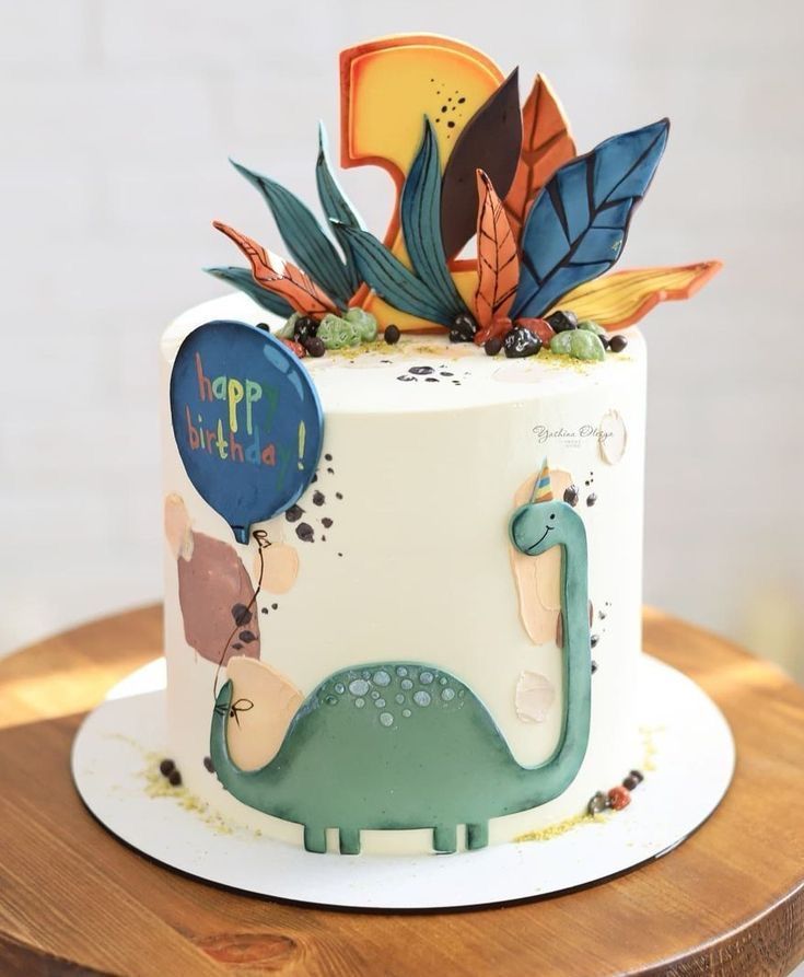 a dinosaur themed birthday cake with balloons and leaves on the top, sitting on a wooden table