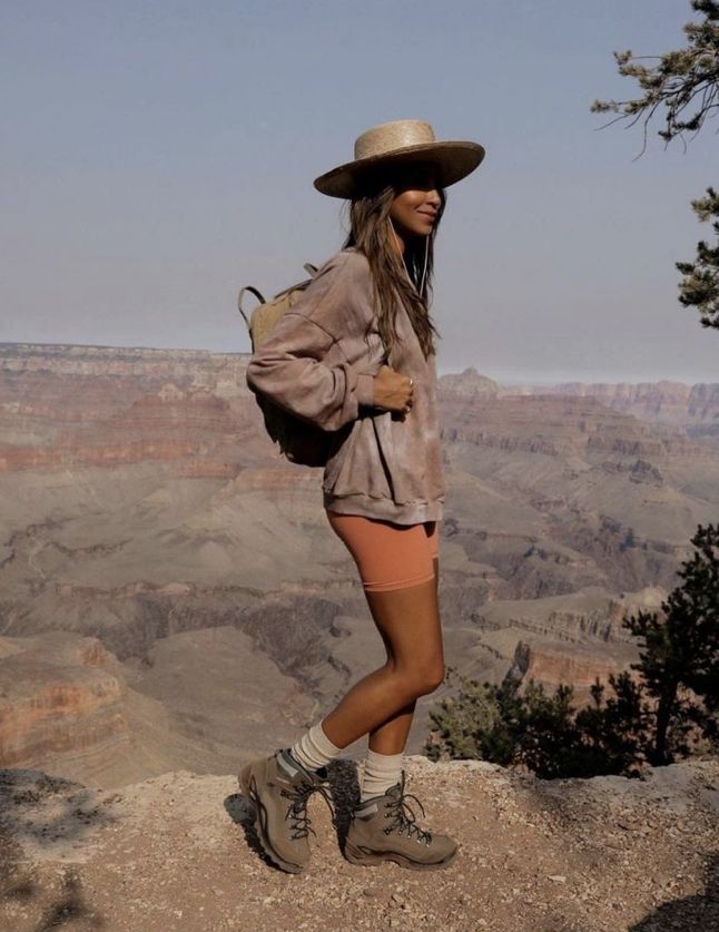 a woman standing on the edge of a cliff wearing a hat and shorts with her legs crossed