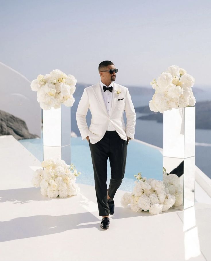 a man in a tuxedo standing next to some white flowers and looking at the camera