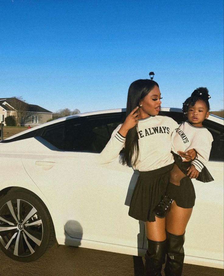 a woman standing in front of a white car holding a small child on her lap