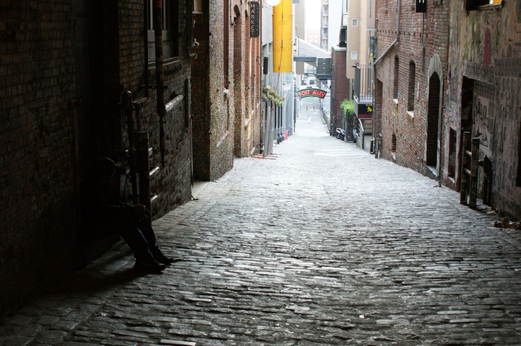 an alleyway with cobblestone floors and tall brick buildings on either side, one person sitting in the doorway