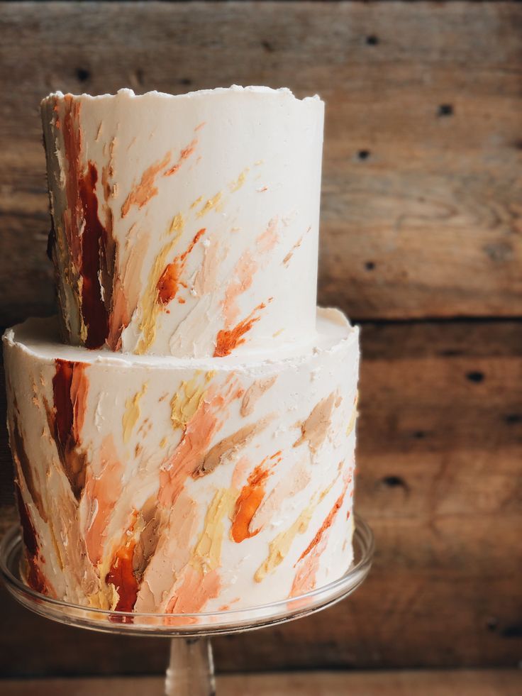 a three tiered cake with orange and white icing on a metal stand against a wooden wall