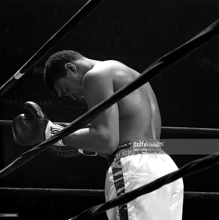 a black and white photo of a man with boxing gloves in his hands, leaning against the ropes