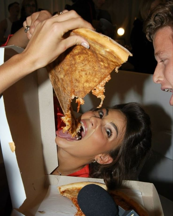 a woman is eating pizza from a box while someone holds her hand over the mouth