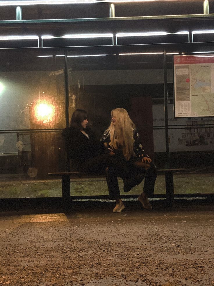 two people sitting on a bench in front of a bus stop at night with the lights on