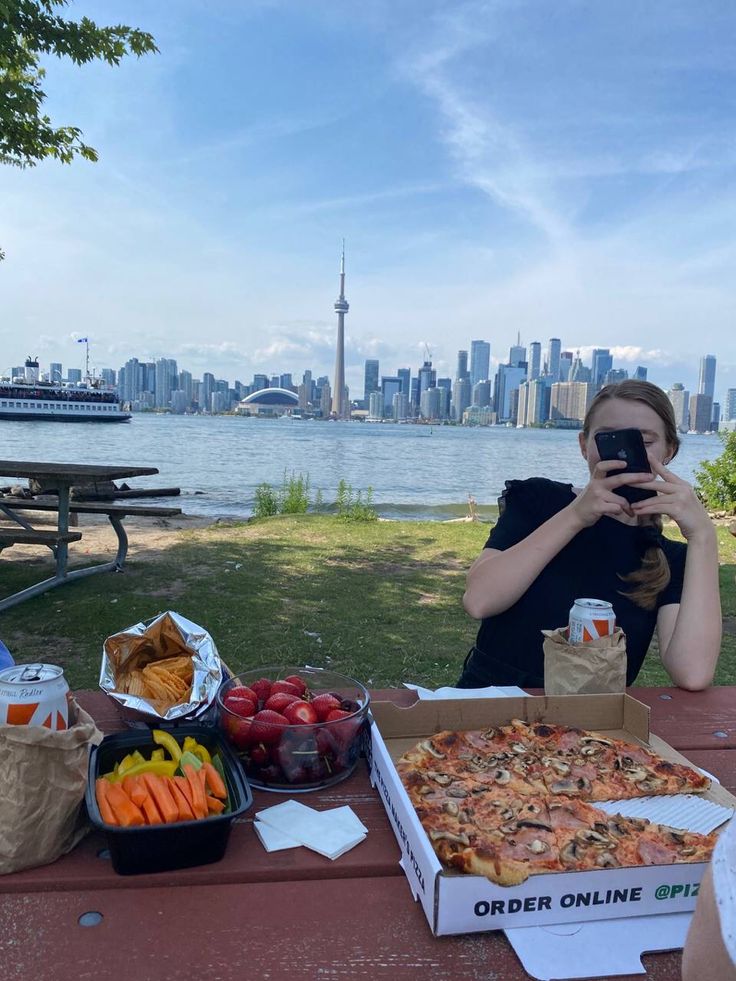 a woman sitting at a picnic table taking a selfie with her cell phone in front of a pizza box