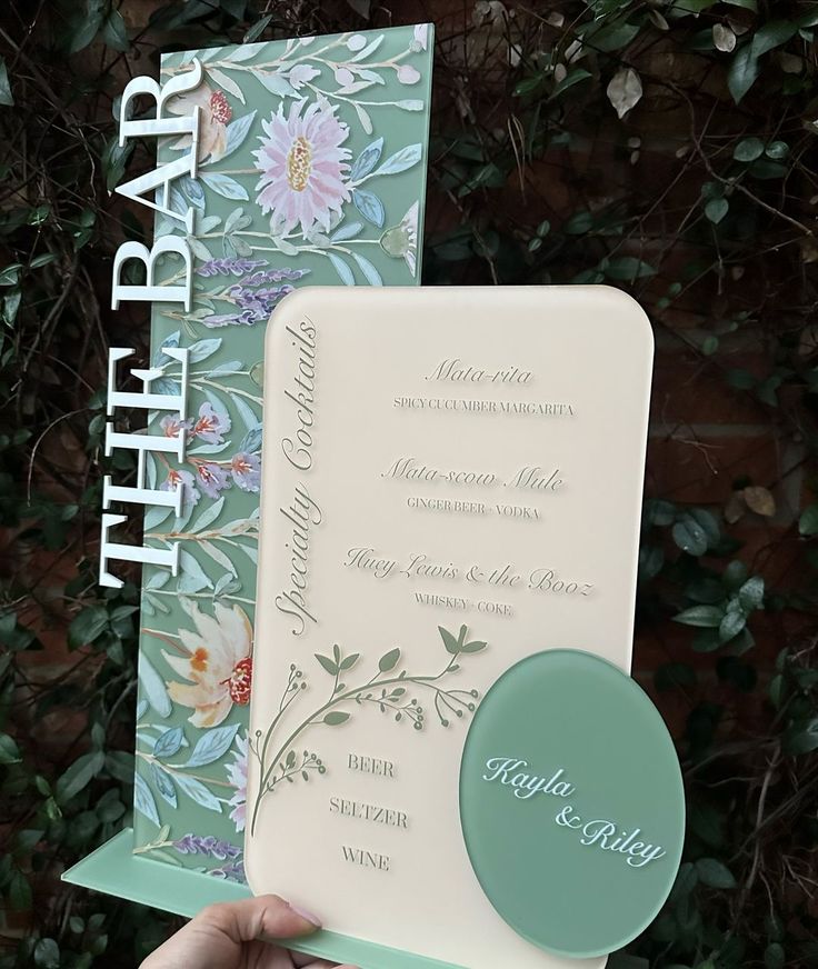 a person holding up a wedding card in front of some flowers and leaves on it