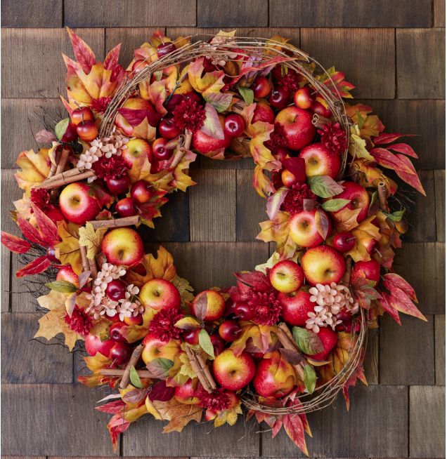 a wreath made out of apples and leaves on a wooden surface with autumn foliage around it