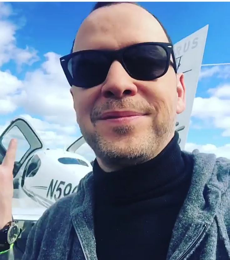 a bald man wearing sunglasses giving the peace sign with his right hand while standing in front of an airplane