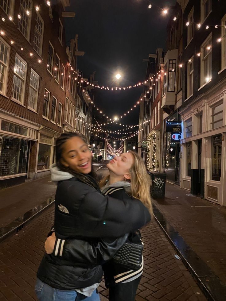 two women hugging each other in the middle of an alleyway at night with lights strung overhead