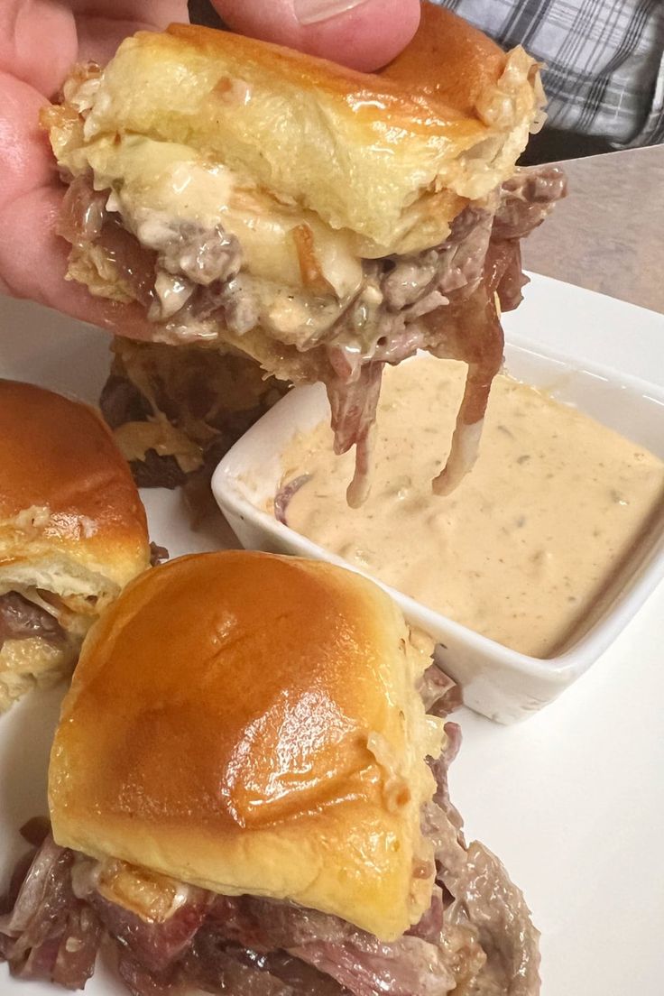 a person holding up a sandwich with meat and cheese on it, next to dipping sauce