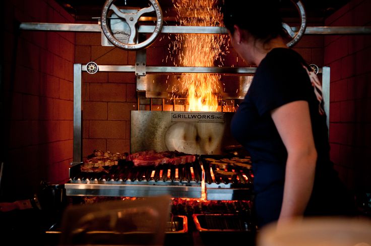 a woman standing in front of a grill filled with meats and hot coals