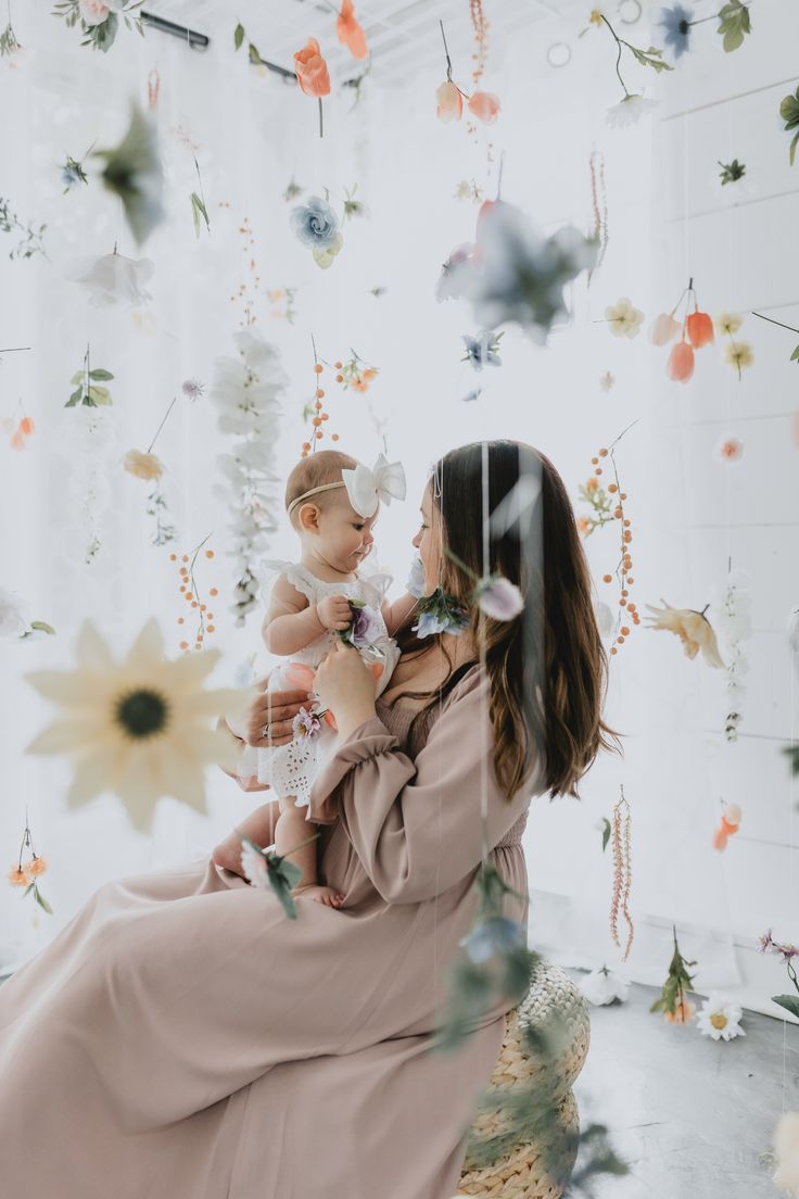 a woman holding a baby in her arms while she looks at flowers hanging from the ceiling