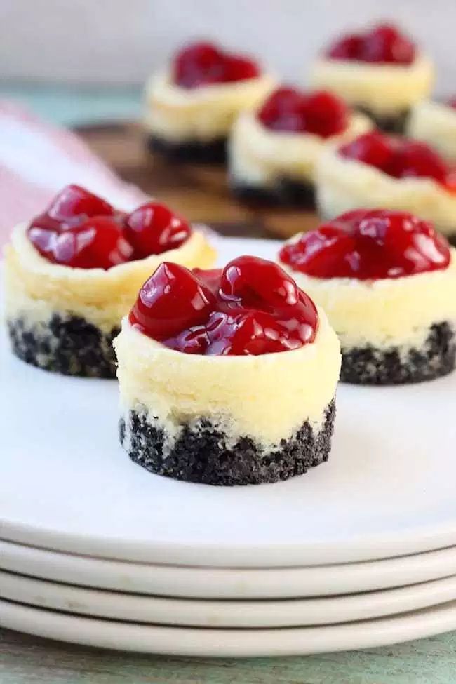 mini cheesecakes with cherries on top are arranged on white plates, ready to be eaten