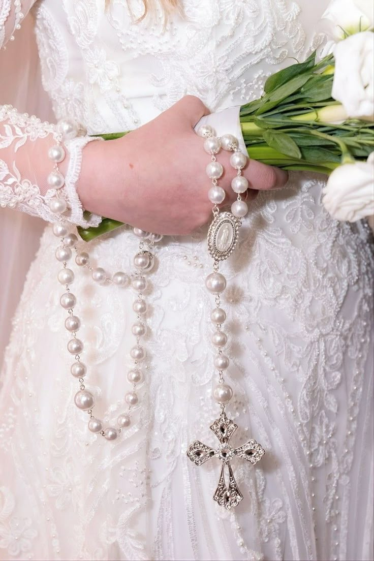 a woman in a wedding dress holding flowers and a cross on her arm with pearls