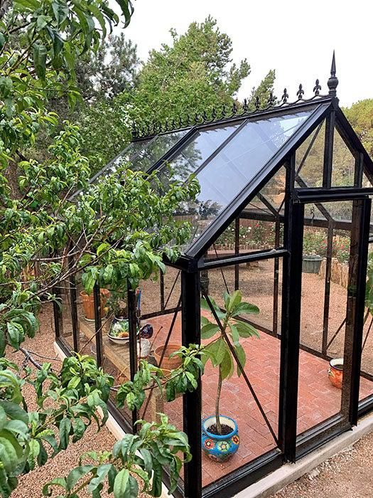 a small black greenhouse in the middle of a yard with potted plants and trees