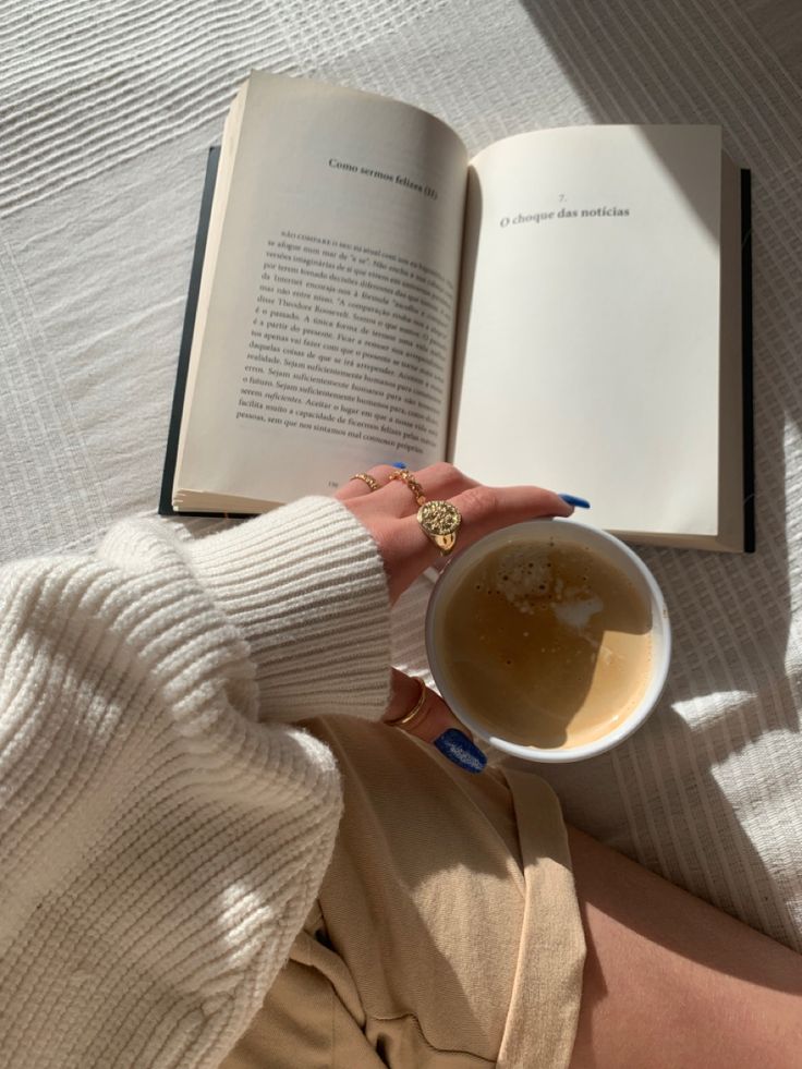 a woman is holding a cup of tea and an open book on her lap while laying in bed