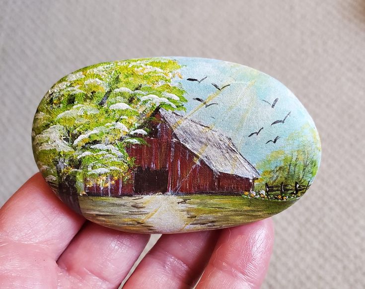 a hand holding a painted rock with a barn and birds flying over the house on it