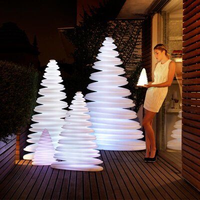 a woman standing next to two white christmas trees on a wooden deck at night time