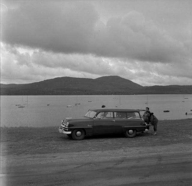 an old black and white photo of a man standing next to a car by the water