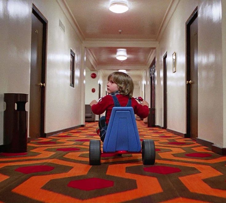 a little boy riding a toy car down a hallway with red and blue carpeting