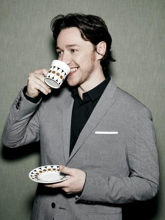 a man in a gray suit drinking from a cup and holding a white plate with black dots on it