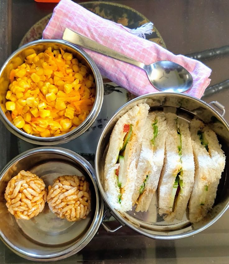 four metal bowls filled with food on top of a glass table next to silver spoons