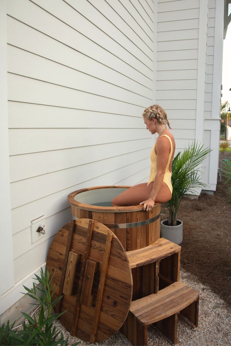 a woman sitting in a wooden tub next to some plants and potted plants on the side of a house