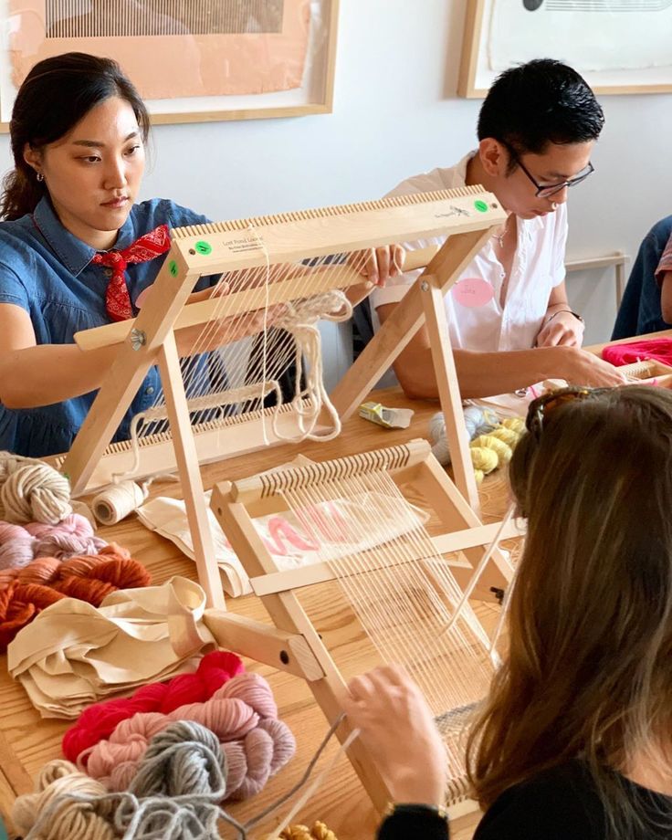 three people sitting at a table working on weavings with yarn in front of them