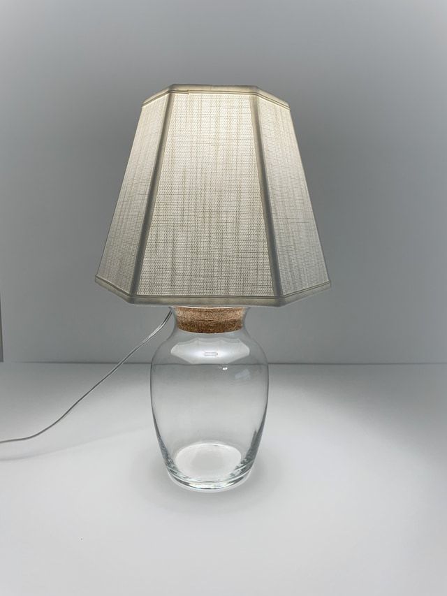 a clear glass lamp with a white shade on it's base and a cord plugged in