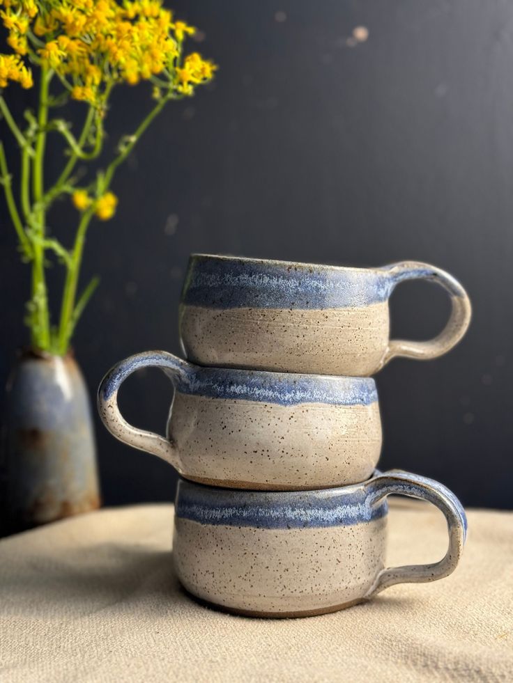 three coffee mugs stacked on top of each other next to a vase with yellow flowers