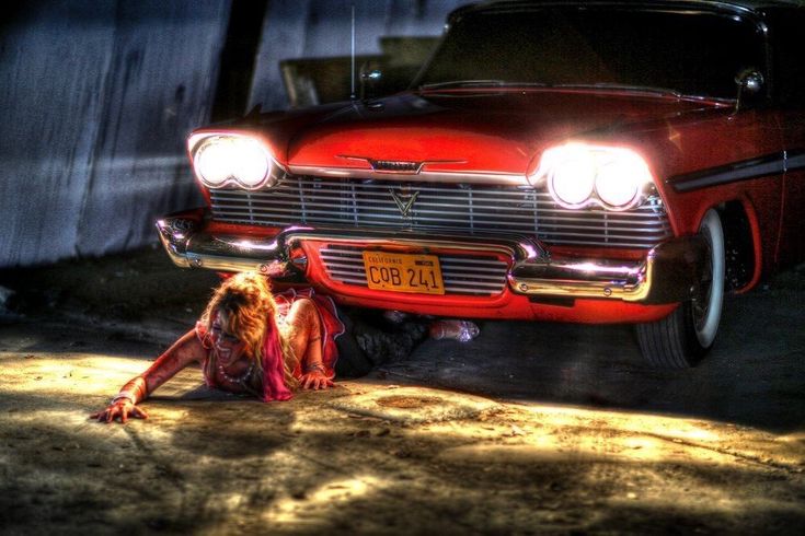 an old red car parked next to a woman laying on the ground in front of it