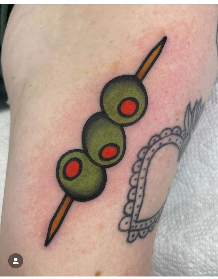 an olive tattoo on the leg of a woman with red eyes and two green olives