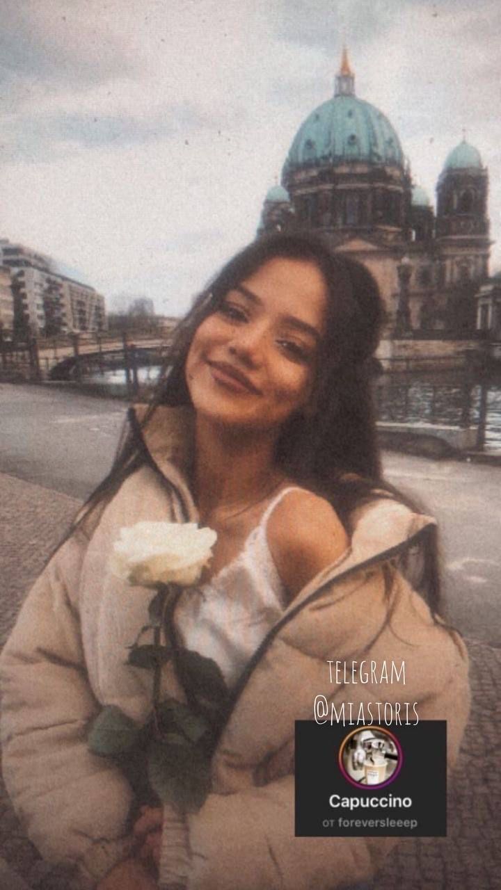 a woman holding a white rose in her right hand and looking at the camera with a building in the background