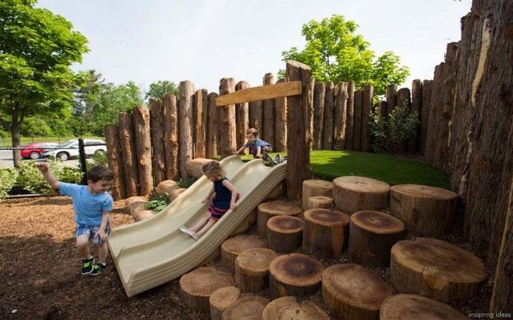 two children playing on a wooden slide in a park with cut down trees and logs