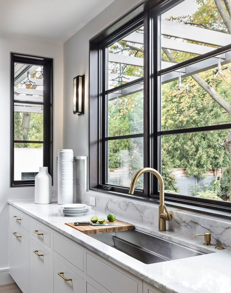 a kitchen with white cabinets and marble counter tops, two large windows above the sink