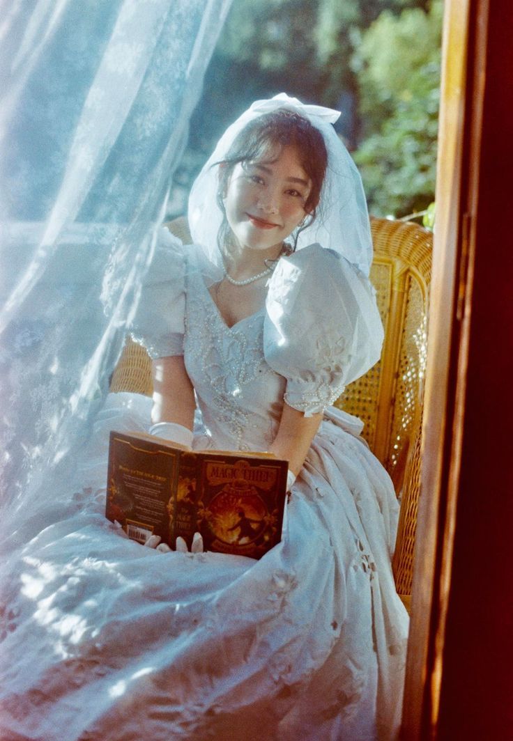 a woman in a white dress sitting on a chair holding a book and wearing a veil