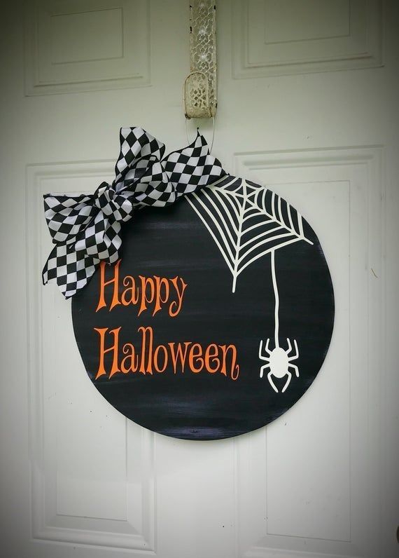 a sign that says happy halloween hanging on the front door with a spider web and checkered bow