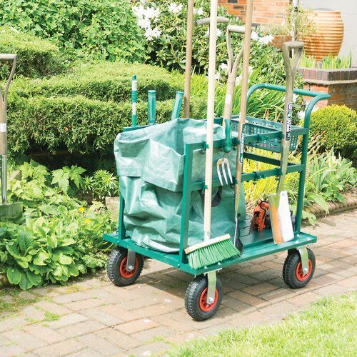 a green hand truck sitting on top of a brick walkway next to bushes and trees