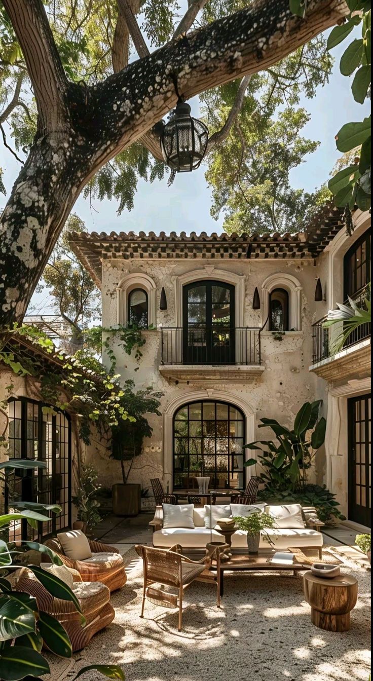 an outdoor living area with couches and tables under a tree in front of a house