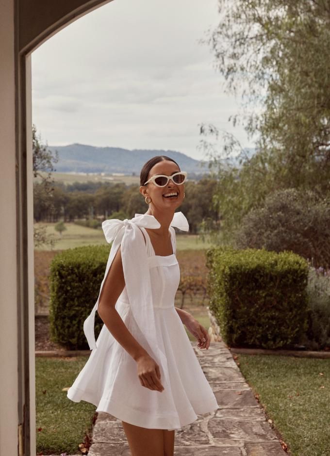 a woman in a white dress and sunglasses walking towards the camera with her hand on her hip